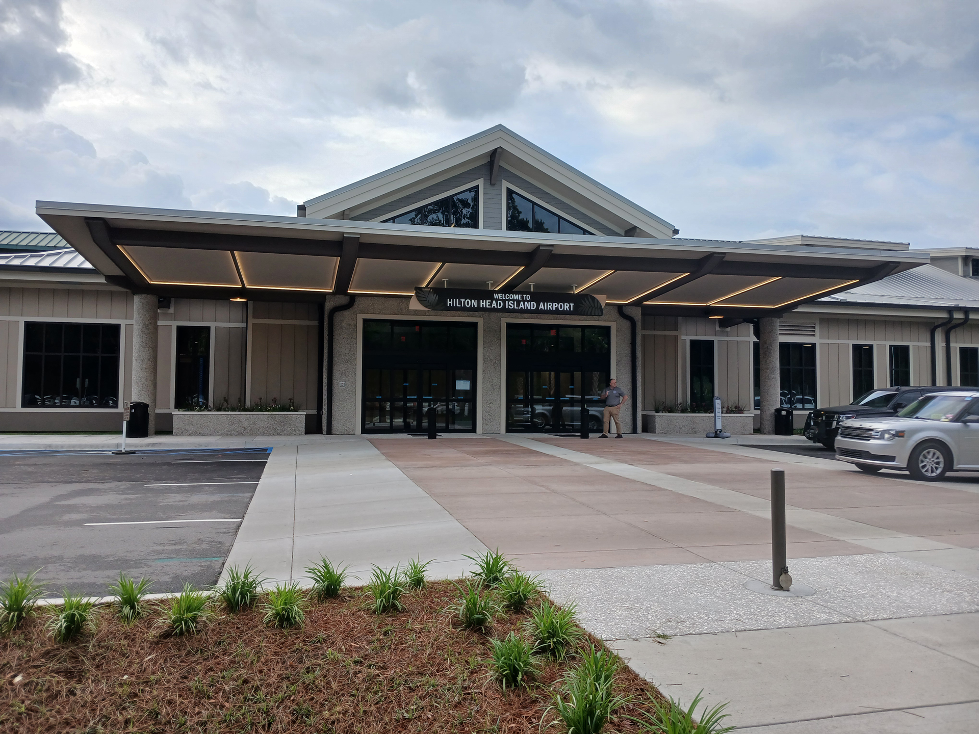 Exterior view of Hilton Head Island Airport with landscaped grounds, palm trees, parking area, and a designated electric vehicle charging space in the foreground.
