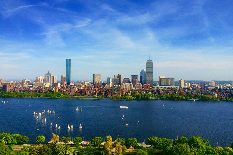 Aerial view of the Boston skyline across the Charles River with sailboats on the water under a clear blue sky.