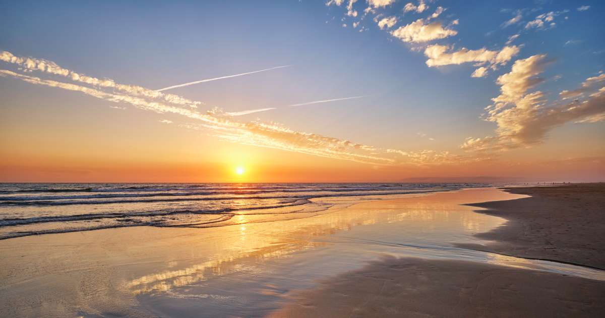 Sunrise over a sandy beach with gentle waves, colorful clouds, and reflections stretching across the wet shoreline.