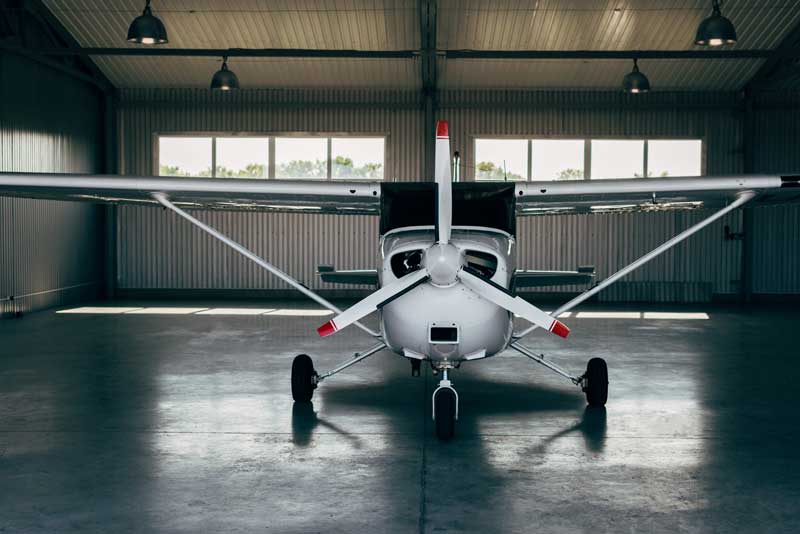Front view of a small single-engine airplane parked inside an aircraft hangar, with natural light coming through the windows behind it.