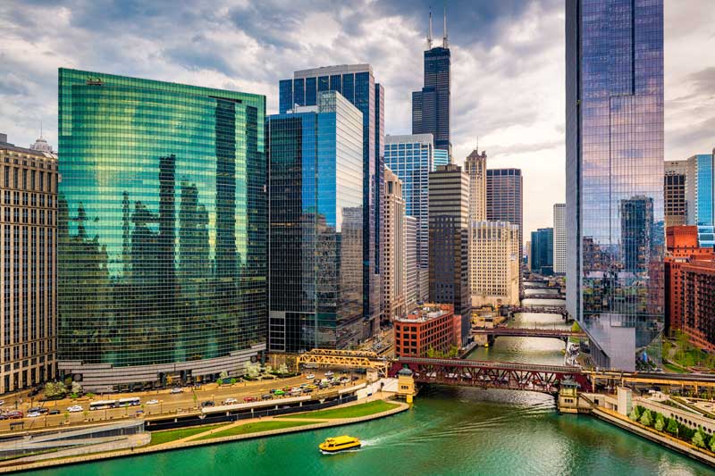 Chicago skyline along the Chicago River, featuring modern glass skyscrapers, a river bridge, and a yellow water taxi under a cloudy sky.