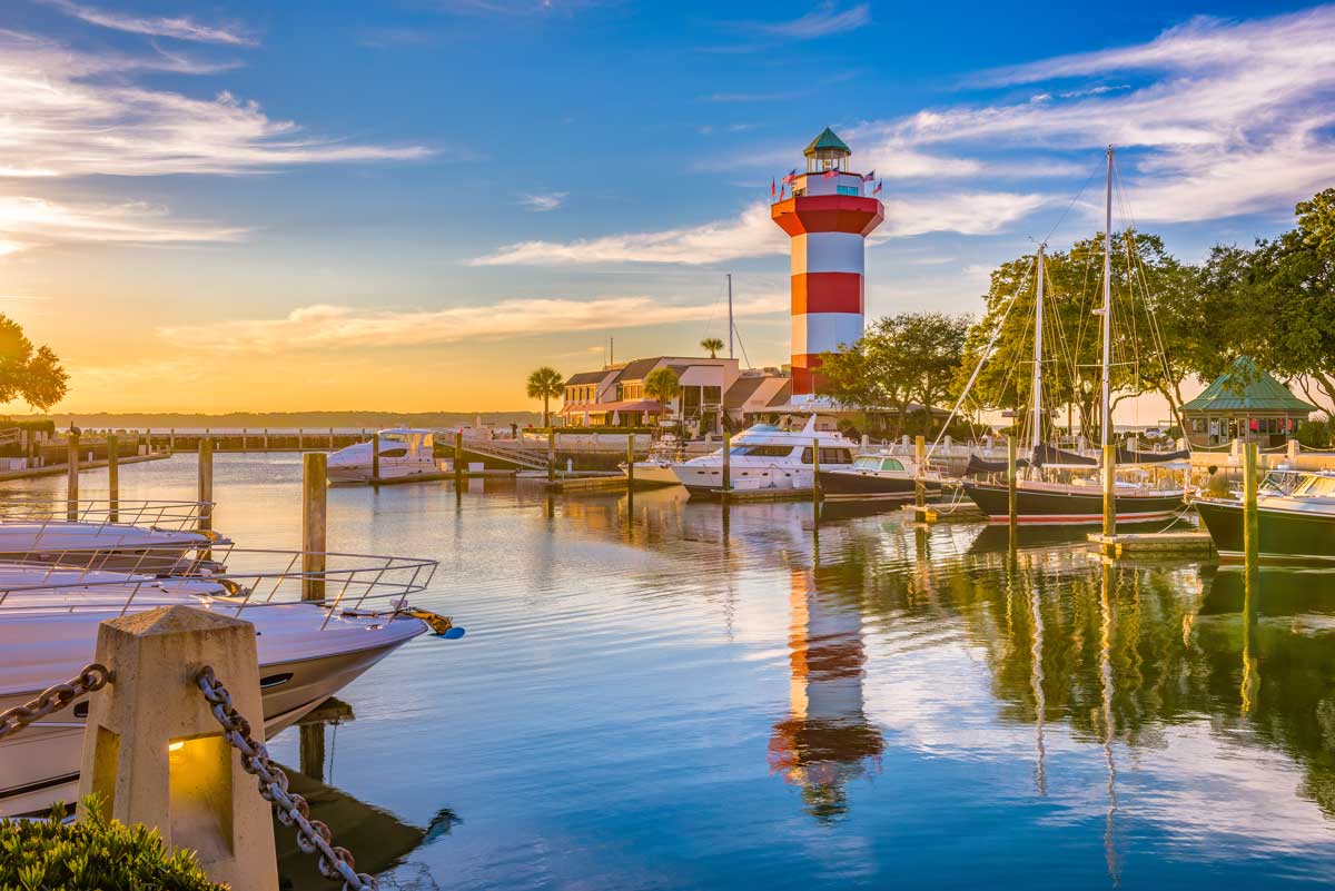 Harbour Town Lighthouse overlooking a marina filled with boats at sunset on Hilton Head Island, with calm water reflecting the colorful lighthouse and sky.