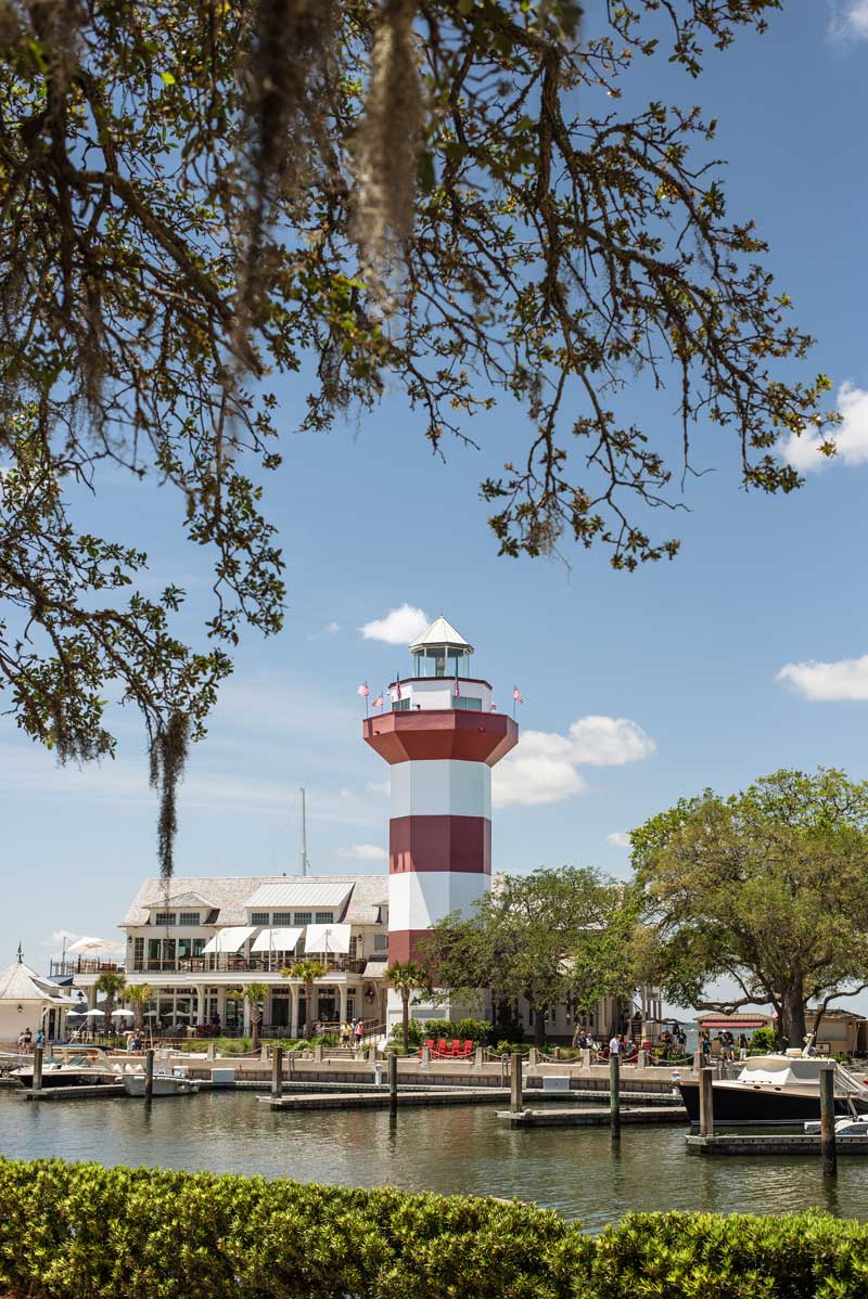 Harbour Town Lighthouse overlooking the marina at Hilton Head Island, framed by oak trees and waterfront docks on a sunny day.