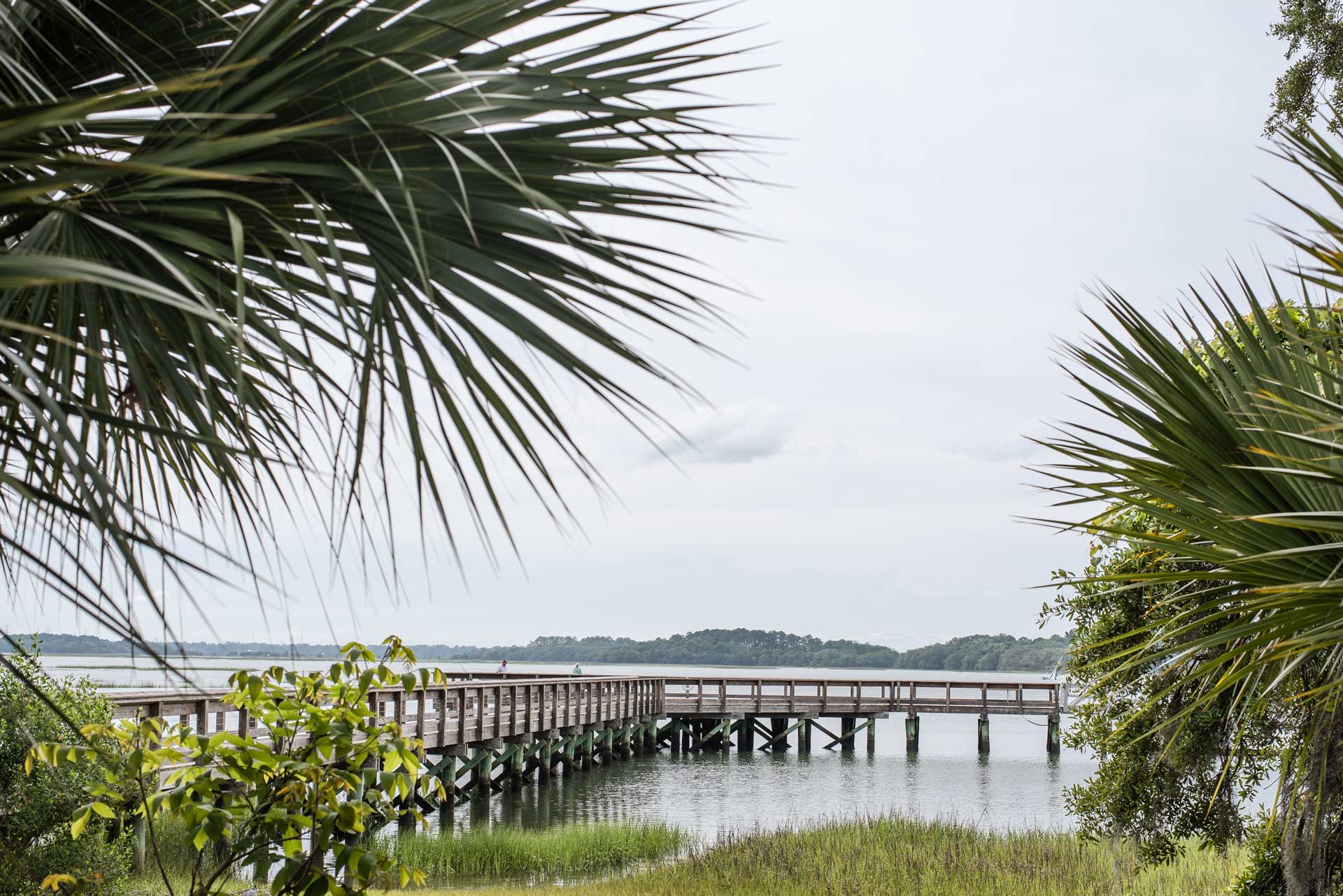 Wooden boardwalk extending over a calm marsh and waterway, framed by palm trees and coastal vegetation under an overcast sky.