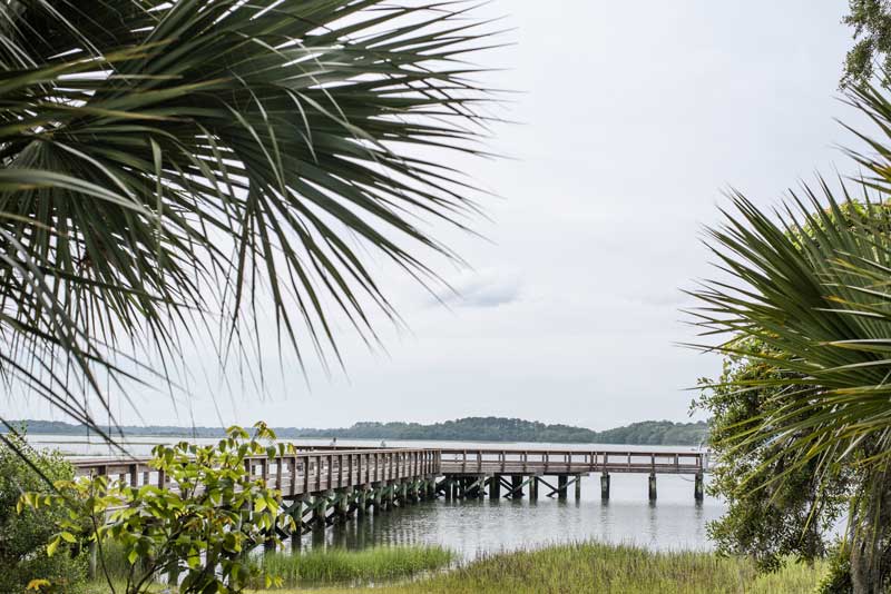 Wooden boardwalk extending over a coastal marsh, framed by palmetto trees and overlooking calm water.