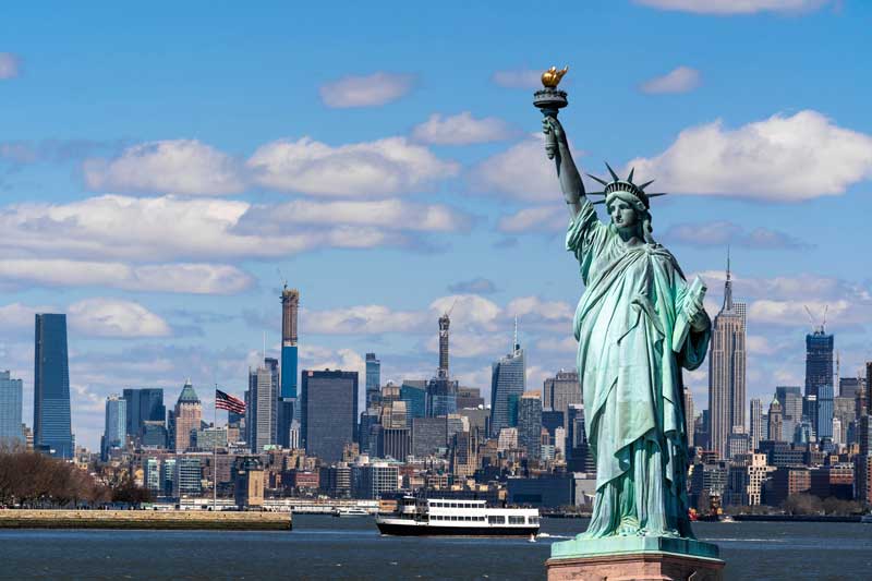 Statue of Liberty standing in New York Harbor with the Manhattan skyline and a ferry boat in the background under a partly cloudy sky.