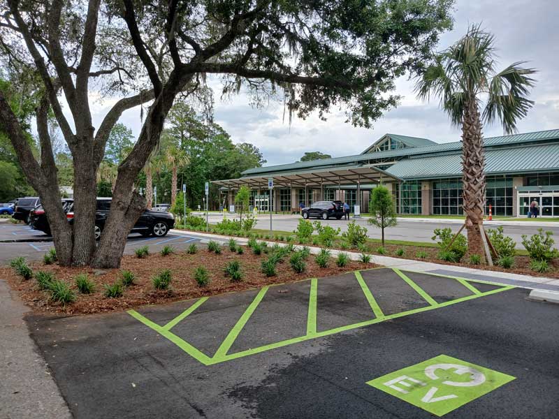 Exterior view of Hilton Head Island Airport with landscaped grounds, palm trees, parking area, and a designated electric vehicle charging space in the foreground.