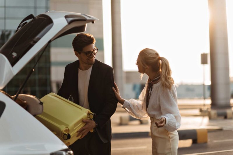 Travelers loading luggage into a vehicle outside an airport terminal during pickup or drop-off.