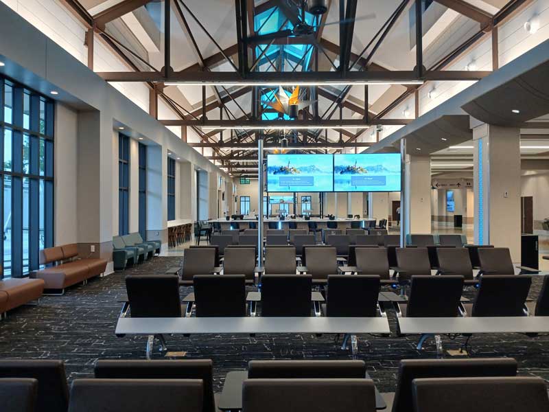Modern airport gate seating area with large windows, overhead flight information displays, and high vaulted ceilings.