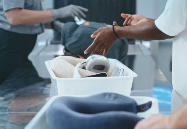 Airport security screening checkpoint with passengers placing belongings in bins while TSA officers inspect carry-on items at the screening area.